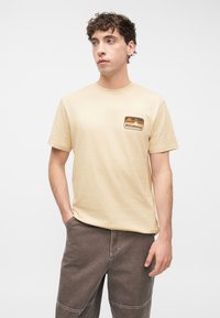 Νεαρός άνδρας με κατσαρά μαλλιά, που φοράει μία μπεζ μπλούζα Billabong και καφέ παντελόνι, στέκεται με το ένα χέρι στην τσέπη μπροστά σε απλό φόντο.