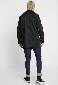 Cappotto nero su misura con vestibilità dritta e maniche lunghe, abbinato a jeans blu scuro e sneaker alte nere. Texture liscia, design minimalista.