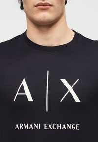 Αρσενικός κορμός που φοράει μαύρο T-shirt Armani Exchange με μεγάλο λευκό λογότυπο A|X και το κείμενο «Armani Exchange» στο στήθος.