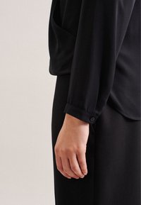 Blusa nera realizzata in tessuto liscio con una vestibilità comoda, caratterizzata da polsini con bottoni e una tasca laterale, abbinata a pantaloni neri coordinati.