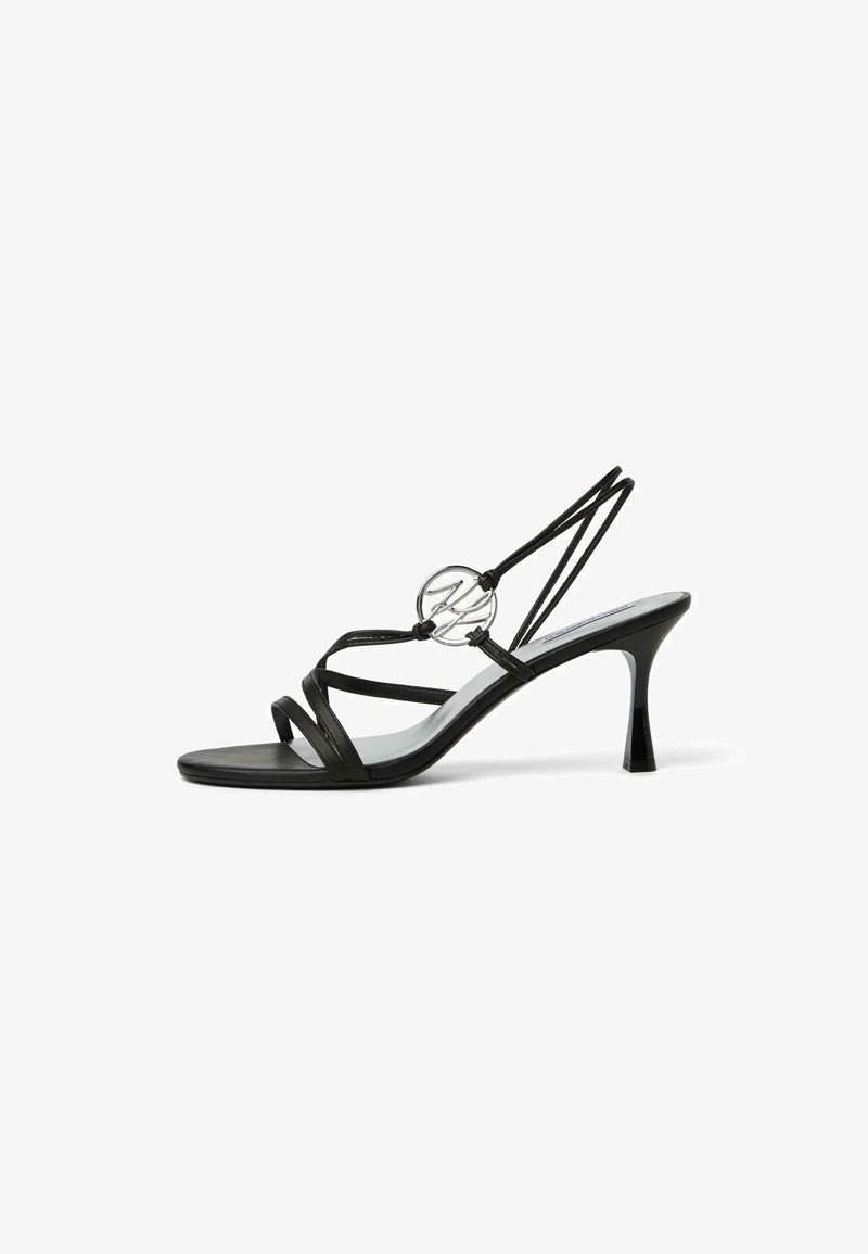Sandale à talons hauts noire à fines brides, avec un détail logo circulaire argenté et un talon effilé sur fond blanc.