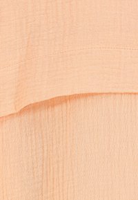 Hellpeachfarbener Stoff mit einer zerknitterten Textur, der horizontale Nähte und ein weiches, fließendes Design aufweist. Dünnes und luftiges Material, das sich gut zum Schichten eignet.