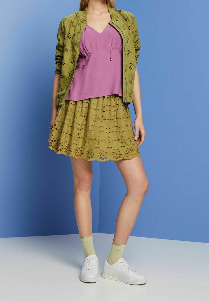 Giacca bomber verde con ritagli floreali su una maglietta viola, abbinata a una gonna verde chiaro con orlo ondulato e sneaker bianche.
