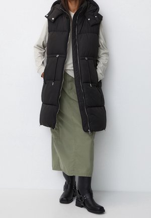 Person wearing a long black padded sleeveless jacket over a beige top, olive green skirt, black tights, and black ankle boots.