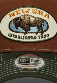 Gorra de béisbol de algodón marrón con visera verde. Presenta un logotipo bordado de bisonte y el texto "NEW ERA". Correa ajustable en la parte posterior.