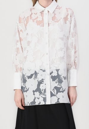 Hvid gennemsigtig bluse med lange ærmer, blomsterblonde-mønster, spids krave, knapper foran og brede manchetter over et sort skørt.