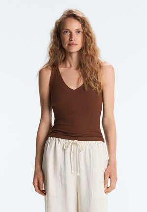 Donna con capelli lunghi e ondulati che indossa un top marrone senza maniche con scollo a V e pantaloni bianchi larghi con coulisse, in piedi contro uno sfondo chiaro e semplice.