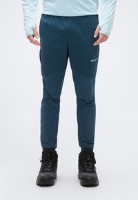 Άτομο που φοράει μπλε σκούρο παντελόνι Columbia για υπαίθριες δραστηριότητες και μαύρα ορειβατικά μποτάκια, στέκεται μπροστά σε ένα απλό λευκό φόντο.