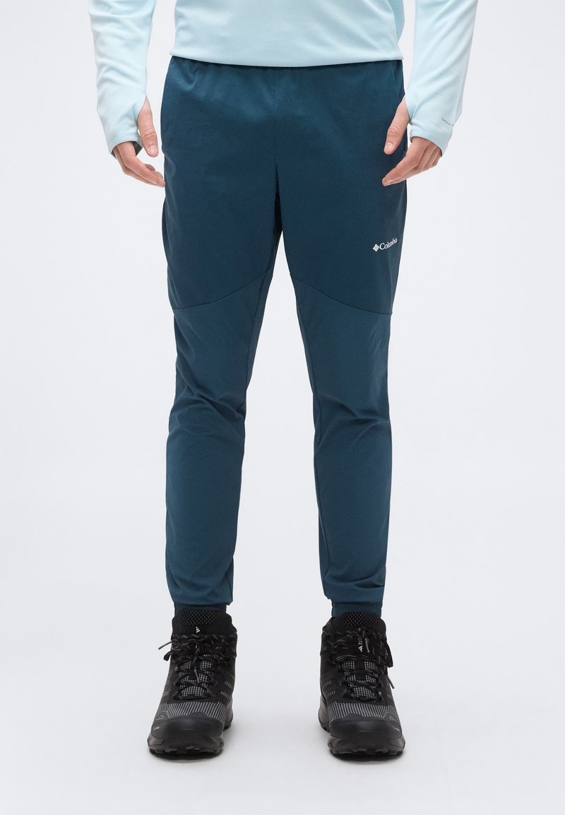 Άτομο που φοράει μπλε σκούρο παντελόνι Columbia για υπαίθριες δραστηριότητες και μαύρα ορειβατικά μποτάκια, στέκεται μπροστά σε ένα απλό λευκό φόντο.