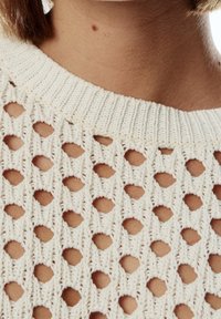 Haut en maille crème présentant un design ajouré avec des motifs en forme de losange et ronds, un col côtelé et une finition texturée.