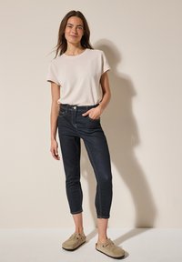 Camisa de manga corta texturizada de color rosa claro, jeans de mezclilla oscura con costuras claras y zapatos beige tipo slip-on. El modelo está de pie contra un fondo neutro.