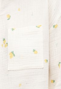 Κοντινή λήψη λευκού υφάσματος με μικρά κίτρινα εκτυπώσεις λεμονιών και ραμμένη τετράγωνη τσέπη με το ίδιο μοτίβο λεμονιών.