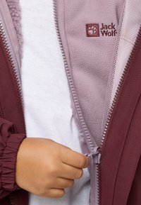 Burgunderjacke mit lila Fleece-Innenfutter und Reißverschluss. Hand greift den Reißverschluss. Weißes Unterhemd ist unter der Jacke sichtbar.