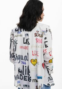 Long-sleeve shirt in white featuring colorful text and graphics, including hearts and animals, with a relaxed fit and buttoned cuffs.