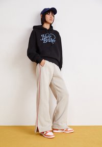 Svart hettegenser med "Heart Breaker" grafikk, beige vide joggebukser med rød side stripe, og oransje og hvite sneakers. Blå caps på hodet.