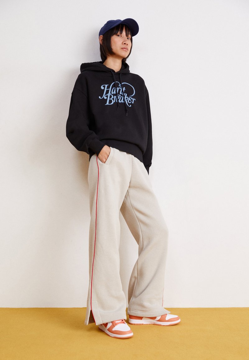 Haut à capuche noir avec un graphique "Heart Breaker", pantalons de survêtement larges beige avec une bande rouge sur le côté, et baskets orange et blanches. Casquette bleue portée.