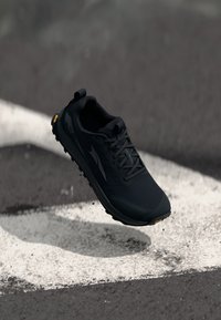Chaussure de sport noire avec un design léger, une tige texturée et une semelle Vibram durable. Équipée de lacets et d'une languette au talon.