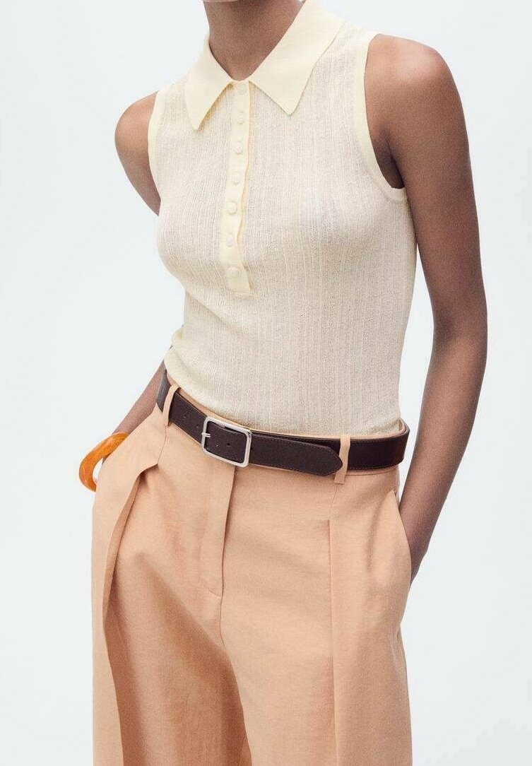 Haut en maille crème sans manches avec patte boutonnée et col, associé à un pantalon beige taille haute et une ceinture marron foncé.