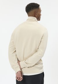 Béžová mikina s vysokým golierom a dlhými rukávmi, s jemnou textúrou a voľným strihom. Zadný pohľad ukazuje minimalistický dizajn.