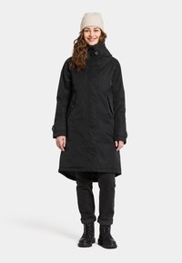 Zwarte waterdichte parka met hoge kraag, zijzakken en een uitlopende zoom. Draag over donkere jeans en zwarte veterschoenen. Beigekleurige gebreide beanie.