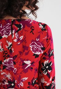 Takapuolet henkilö, jolla on kiharat hiukset, pukeutuneena punaiseen paitaan, jossa on violetteja, mustia ja valkoisia kukka- ja perhoskuvioita.