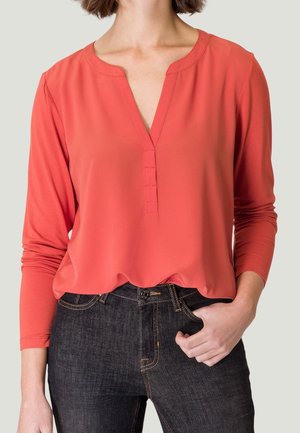 Vrouw draagt een koraalroze blouse met lange mouwen en V-hals met knoopdetails, gecombineerd met donkere spijkerbroek, hand in de zak.