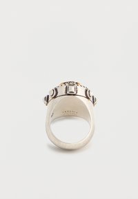 Anillo Versace de tono plateado con detalles decorativos en relieve y logotipo grabado en el interior de la banda, mostrado sobre un fondo blanco.