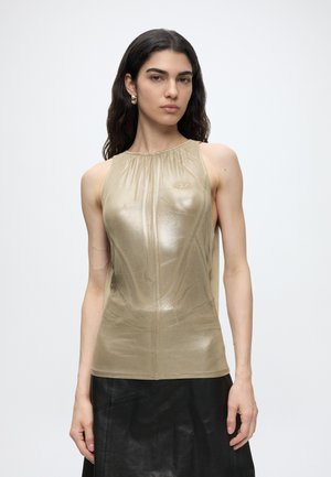 Femme aux longs cheveux foncés portant un top beige brillant sans manches et une jupe noire, debout devant un fond clair uni.