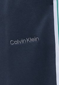 Ναυτικό ύφασμα με γκρι κεντητό λογότυπο «Calvin Klein» και μια λευκή κάθετη ρίγα με πράσινη γραμμή κατά μήκος της δεξιάς άκρης.
