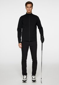 Svart zip-up jacka gjord av ett texturerat material, figursydd design, i kombination med svarta byxor och sportskor med vita accenter, som håller en golfklubba.