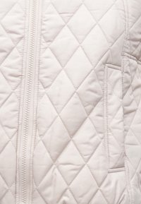 Jasno beżowa pikowana kurtka z przeszyciami w kształcie diamentów, centralnym zamkiem błyskawicznym oraz boczną kieszenią o teksturowanym wykończeniu.