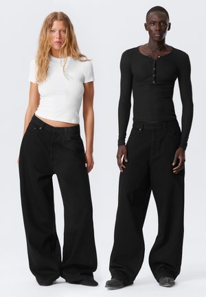 Schwarze weite Jeans und figurbetonte Langarmoberteile, eines weiß und eines schwarz, präsentiert auf zwei Modellen vor einem neutralen Hintergrund.