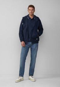 Man staat gekleed in een marineblauw zip-up jack, een marineblauwe polo, lichtblauwe spijkerbroek en witte sneakers tegen een effen grijze achtergrond.