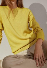 Femme assise contre un mur portant un pull jaune vif et un pantalon beige, avec une bague en or à la main droite reposant sur son genou.