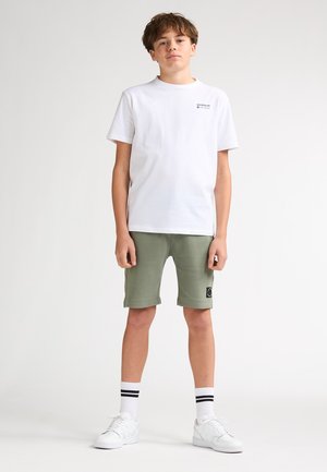 Camiseta blanca de algodón con un pequeño logo, pantalones cortos verdes con dobladillos enrollados y zapatillas blancas con detalles negros. Calcetines negros con dos rayas blancas.