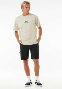 Beige bomuld t-shirt med blå logo, kombineret med sorte shorts og sorte sneakers. Outfittet har en afslappet pasform og casual stil.