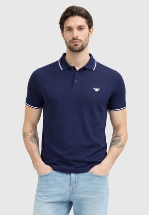 Mann mit kurzem dunklem Haar und Bart, der ein marineblaues Polohemd mit weißem Besatz und hellblaue Jeans trägt, steht mit den Händen in den Taschen.