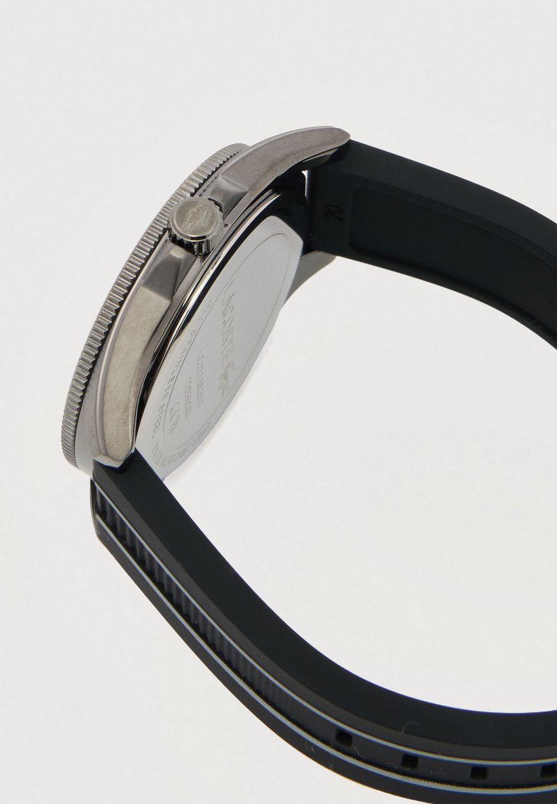 Zijaanzicht van een horloge met een zilveren metalen kast, een gestructureerde kroon en een zwarte rubberen band tegen een effen achtergrond.