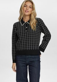 Pull en tricot noir avec un motif en damier, accents blancs, col boutonné et poignets côtelés. Associé à un jean en denim bleu foncé.