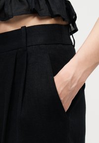 Άτομο που φοράει μαύρο παντελόνι με ψηλή μέση και έχει το χέρι στην τσέπη, καθώς και κοντό μαύρο τοπ, με εκτεθειμένη τη μέση, μπροστά σε απλό φόντο.