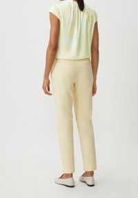 Femme portant un haut à manches courtes à motifs jaune clair, un pantalon jaune pâle et des mocassins beiges, de dos face au spectateur.