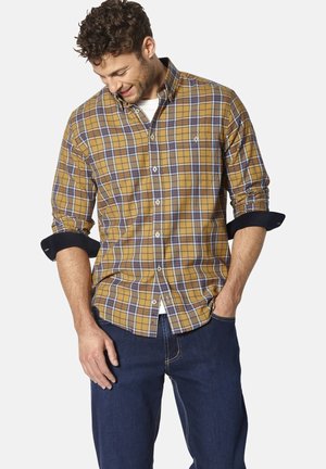 Langärmliges kariertes Hemd in Gelb, Blau und Weiß; mit einem Button-down-Kragen, hochgerollten Ärmeln und einer Brusttasche. Trägt dazu eine dunkle Jeans.