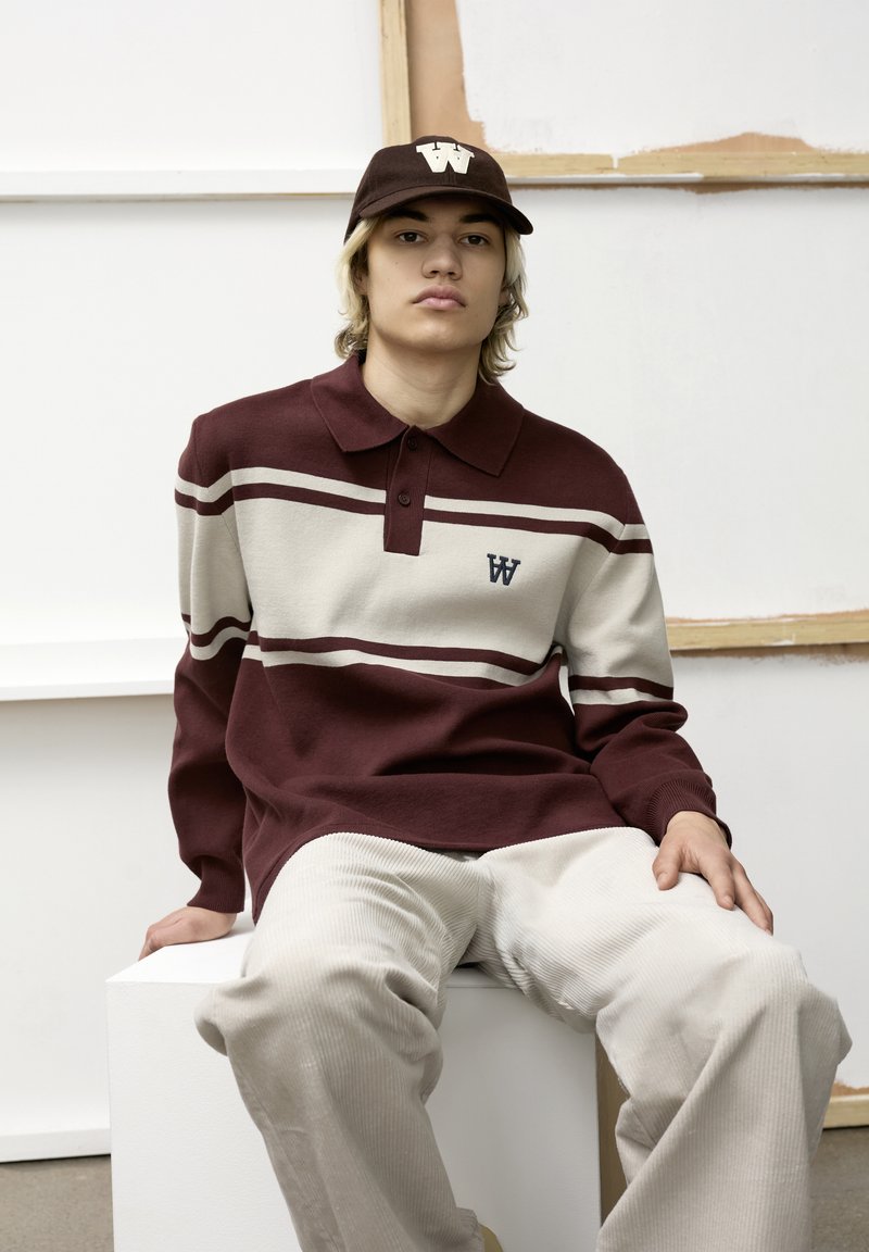 Ung person med lyst hår iført en bordeauxfarvet og hvid stribet sweater og matchende kasket, siddende på en hvid kube mod en ensfarvet baggrund.