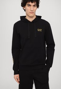 Black hoodie made of soft fabric, featuring a front pocket, drawstring hood, and gold EA7 logo on the chest and sleeve. Long-sleeved design.