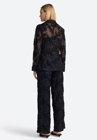 Mustlane ülikond, mis sisaldab läbipaistvat, tikandiga jakki lillemustrite ja sädemetega, millega on kombineeritud tekstuuriga laiad püksid.