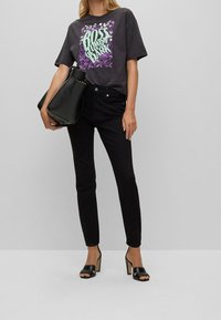 Žena v černém grafickém tričku, černých skinny džínách a černých sandálech na vysokém podpatku, držící velkou černou kabelku na šedém pozadí.