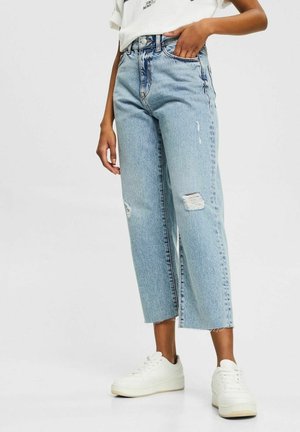 Person trägt hellblaue, kurz geschnittene, zerrissene Jeans, weiße Turnschuhe und ein weißes, in die Hose gestecktes T-Shirt und steht mit einer Hand in der Tasche.