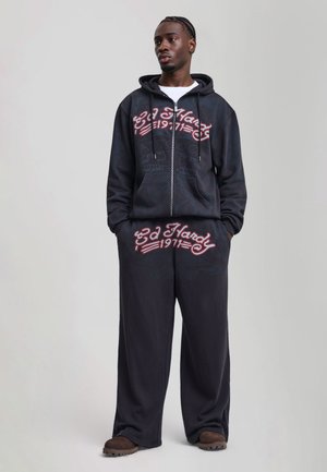 Uomo che indossa una felpa con cappuccio nera Ed Hardy e pantaloni della tuta abbinati con logo rosso, in piedi con le mani nelle tasche su uno sfondo semplice.
