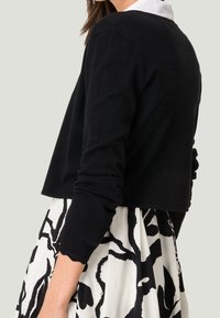 Zwarte cropped sweater van zacht materiaal met lange mouwen, gedragen over een wit en zwart opgemaakt rok. Heeft een ronde halslijn en minimale details.