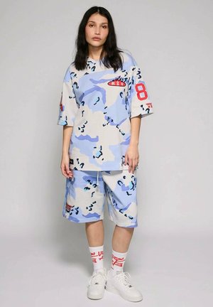 Frau trägt ein übergroßes Hemd mit hellblau-weißem Tarnmuster und passende Shorts, weiße Socken mit rotem Aufdruck und weiße Turnschuhe.